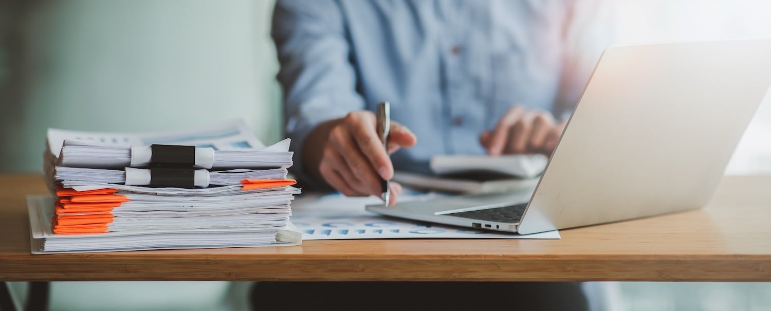 A person writing at a desk with a stack of clipped documents and an open laptop.