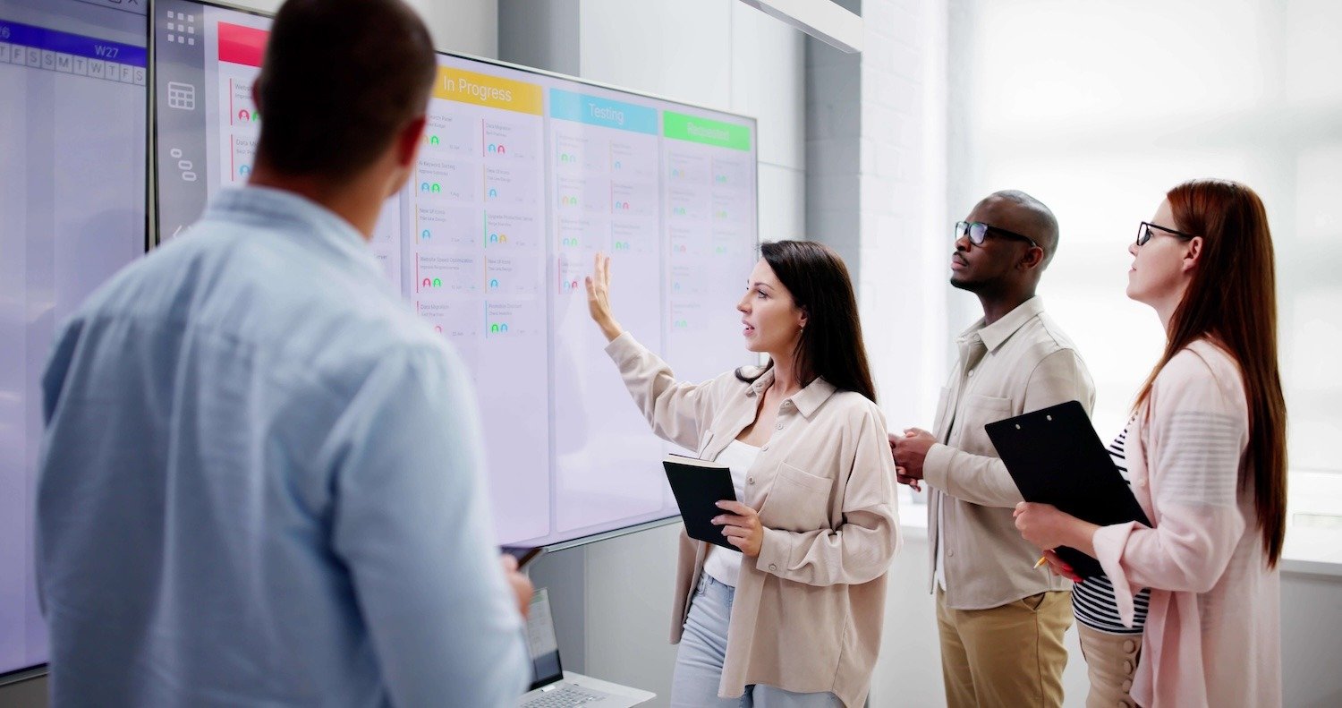 Four people in a modern office look at a large digital screen displaying a chart.